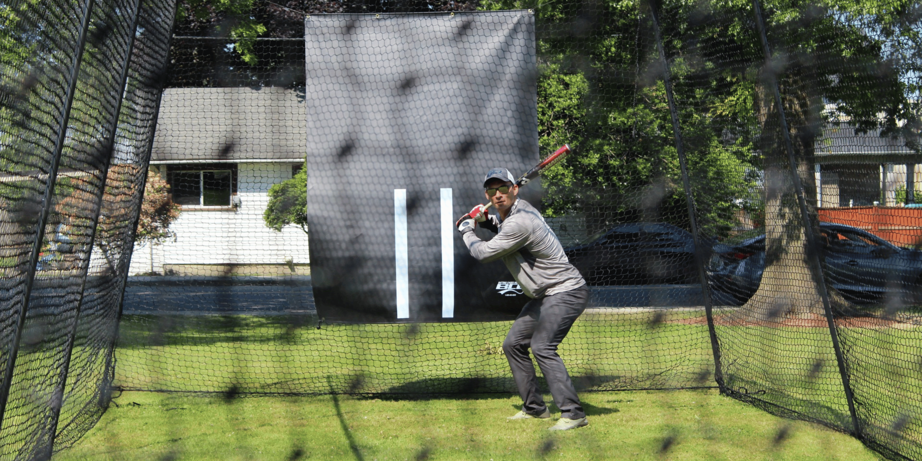 A person stands in a batting cage outdoors, holding a baseball bat in a ready stance with a target and netting in the background.