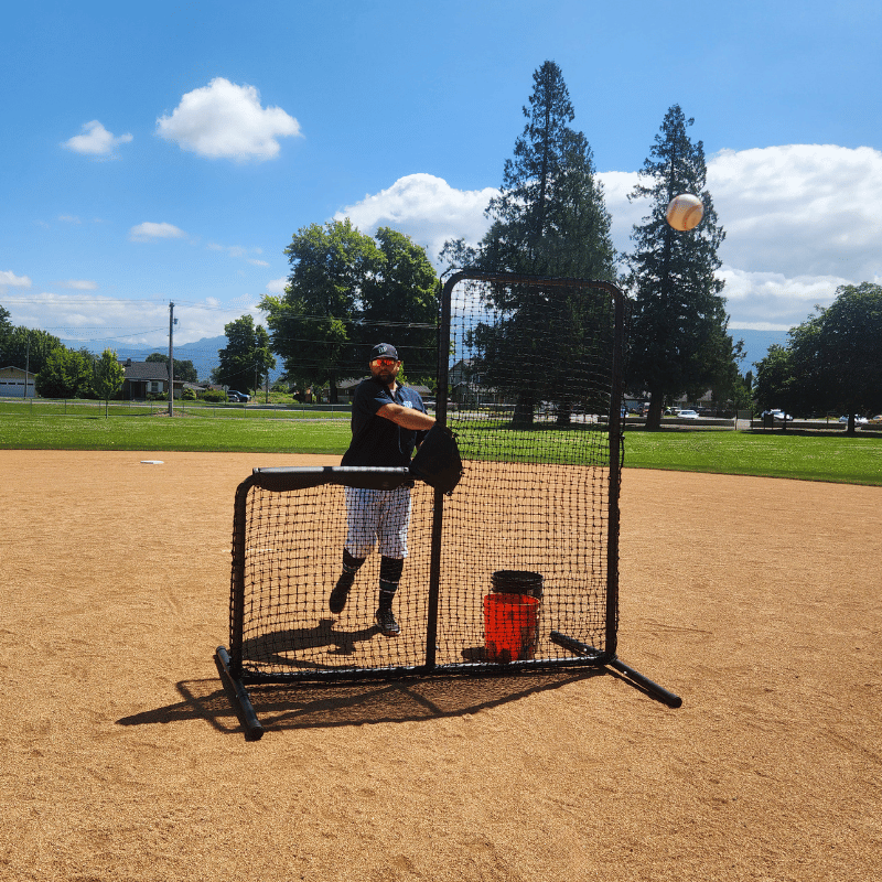 A baseball field featuring the BATTING CAGES INC Armadillo Protective L-Screen (Aluminum) and a black bucket on the pitcher’s mound under a clear sky.