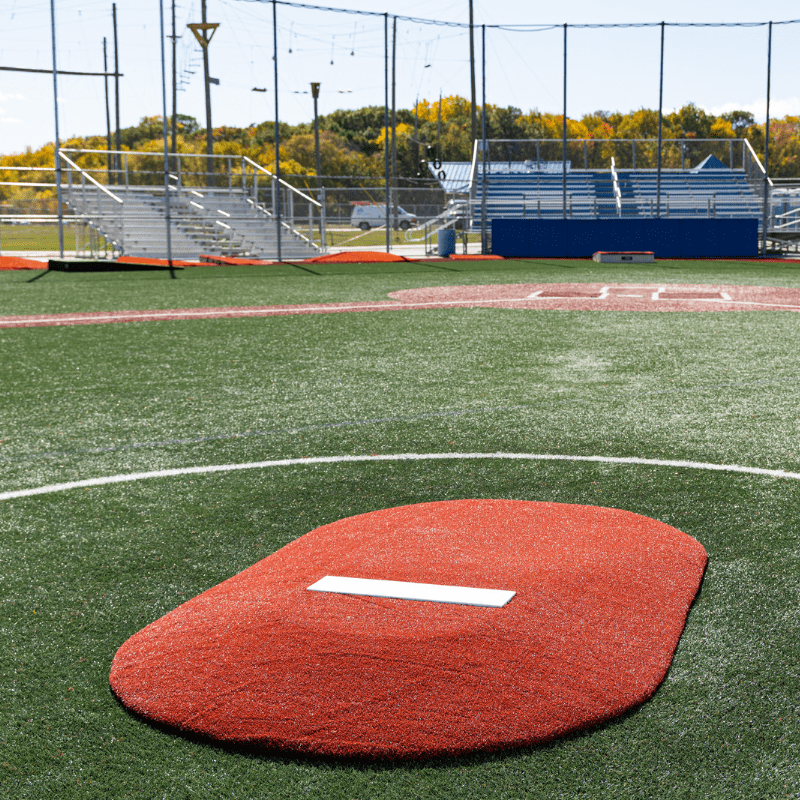 A synthetic turf baseball field features the PORTOLITE Portolite 6in Full Length Game Mound with a red top and white pitching rubber, empty bleachers, and a safety net in the background.