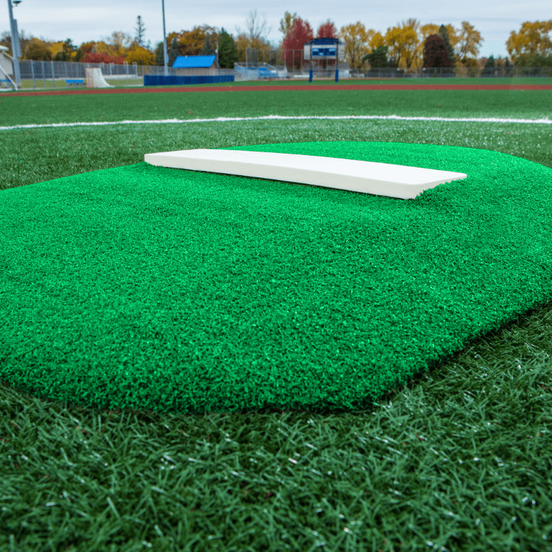 A child in red athletic shoes and striped pants stands on a PORTOLITE 4" Economy Youth Mound, surrounded by green artificial turf.