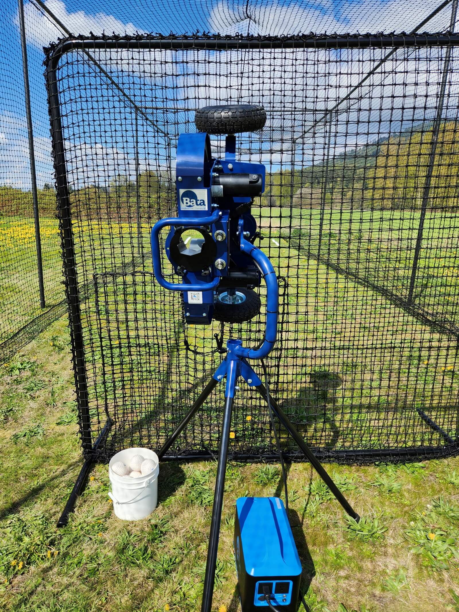 Bata 2 pitching machine with balls and battery behind pitching machine screen in the Thumper Batting cage 